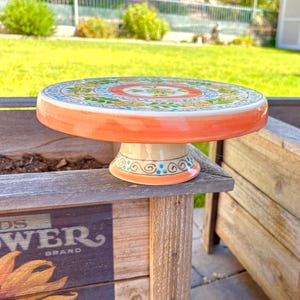 May include: A ceramic cake stand with a round, decorated top and a pedestal base. The top is adorned with a floral pattern in shades of yellow, blue, and green, with an orange border. The base is cream-colored with blue and orange accents.