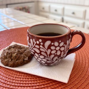 May include: A dark red ceramic mug filled with black coffee, featuring a white floral and leaf pattern painted on the exterior. Beside the mug sits a round, textured brown cookie on a white paper napkin, creating a simple and clear composition.