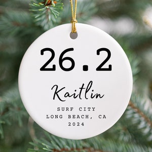 May include: A white ceramic Christmas ornament with the text "26.2" and the name "Kaitlin" in black. Below the name are the words "SURF CITY LONG BEACH, CA 2024". The ornament hangs from a gold string against a backdrop of a Christmas tree.