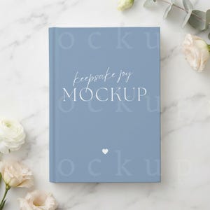 May include: A dusty blue hardcover book with the text "keepsake joy" in a white script font above the word "MOCKUP" in a white serif font, featuring a small white heart icon centered below the text on the front cover of the book.