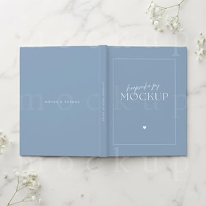 Puede incluir: Un libro de tapa dura azul claro con el texto NOTES & THINGS en la cubierta izquierda y keepsake joy MOCKUP con un pequeño icono de corazón en la cubierta derecha. El libro se muestra en una posición abierta y plana con un acabado sólido.
