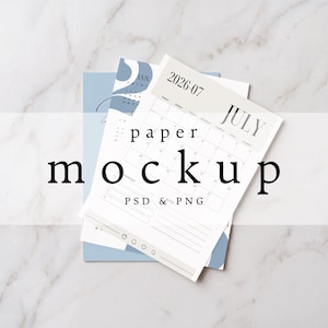May include: A paper mockup featuring a July 2026 calendar page with a light beige header and a blue background sheet. The calendar includes a grid for dates, a priorities section, and small icons at the bottom. The text reads 2026-07, JULY, paper, mockup, PSD & PNG.