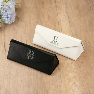 May include: Two eyeglass cases, one black and one white, are displayed on a wooden surface. The black case has the name "Baron" and the letter "D" printed on it. The white case has the name "Evadne" and the letter "E" printed on it.