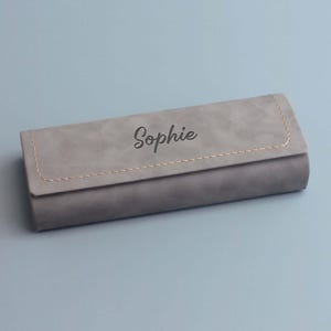 Peut inclure: Un &eacute;tui &agrave; lunettes rectangulaire, gris, doux, avec une couture cousue et le nom "Sophie" grav&eacute; sur le dessus.