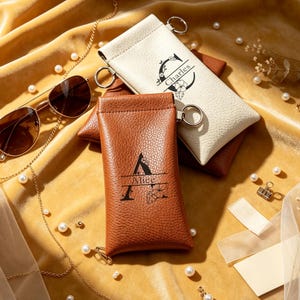 May include: Two rectangular, textured pouches with metal rings, one cream and one tan. The cream pouch features a black letter C and the name Charles with floral accents. The tan pouch displays a black letter A and the name Alice with floral accents.