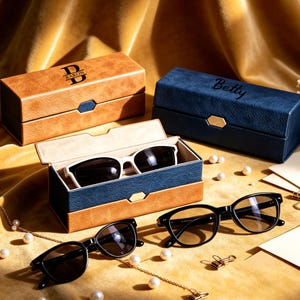 May include: Two rectangular spectacle cases with textured surfaces, one tan with the text BARON and one blue with the text Betty. One open blue case contains white-framed sunglasses. Two pairs of black-framed sunglasses rest on the surface.