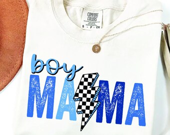 May include: Cream-colored t-shirt with the words "boy mama" in blue and a black and white checkered lightning bolt graphic. A gold necklace with a pendant is on the shirt. A brown hat is visible in the background.