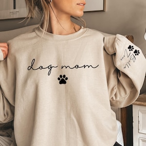 May include: A beige sweatshirt with the words "dog mom" in black script and a black paw print. The sleeve features paw prints and the name "Bella" in script. The sweatshirt is a casual, comfortable style.