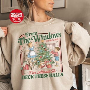 May include: Beige crewneck sweatshirt with a Christmas-themed graphic. The design features a vintage illustration of children decorating a Christmas tree, with text that reads "From The Windows To The Walls, I'm About To Deck These Halls."