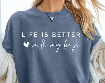 May include: A blue t-shirt with the text "LIFE IS BETTER with my boys" in white script. The shirt is worn by a person with sunglasses and a white sweater. The person is holding a glass of lemonade.