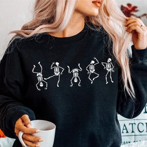 May include: Black sweatshirt featuring a white graphic of dancing skeletons. The crew neck sweatshirt is made of a soft material. The image also includes a white mug and a decorative pillow with the word "KIN" on it.