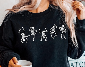 May include: Black sweatshirt featuring a white graphic of dancing skeletons. The crew neck sweatshirt is made of a soft material. The image also includes a white mug and a decorative pillow with the word "KIN" on it.