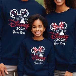 Puede incluir: Sudaderas azul marino con un dise&ntilde;o de Disney. El dise&ntilde;o incluye orejas de Mickey Mouse, copos de nieve, un castillo y el texto "Disney! 2026 Your Text" en rojo y blanco. Las sudaderas son usadas por una familia.