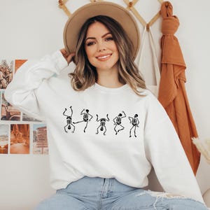 May include: White sweatshirt featuring a black graphic of dancing skeletons. The long-sleeved top is made of a soft material and is suitable for casual wear. The design is a fun, spooky theme.