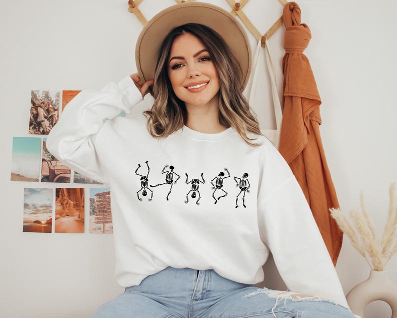 May include: White sweatshirt featuring a black graphic of dancing skeletons. The long-sleeved top is made of a soft material and is suitable for casual wear. The design is a fun, spooky theme.
