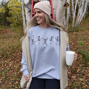 May include: A light gray sweatshirt with a black skeleton dance design. The person is wearing a beige beanie and holding a white mug. The background includes trees and fall foliage.