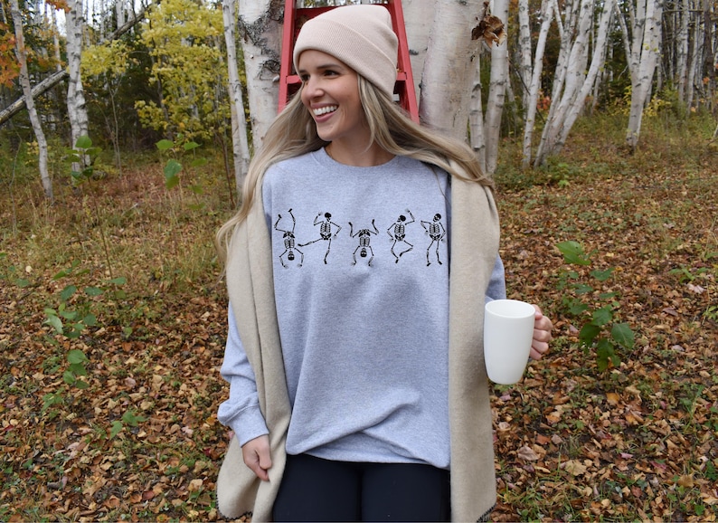 May include: A light gray sweatshirt with a black skeleton dance design. The person is wearing a beige beanie and holding a white mug. The background includes trees and fall foliage.