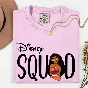 May include: A light pink Comfort Colors t-shirt with the words "Disney Squad" in black script and a cartoon illustration. A gold star pendant necklace and a brown hat are also in the image.