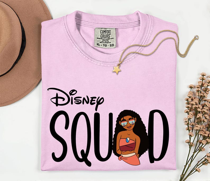 May include: A light pink Comfort Colors t-shirt with the words "Disney Squad" in black script and a cartoon illustration. A gold star pendant necklace and a brown hat are also in the image.
