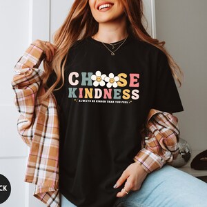 Peut inclure: T-shirt noir avec l'inscription "CHOOSE KINDNESS" dans un style r&eacute;tro color&eacute;. Le motif comprend des marguerites et la phrase "ALWAYS BE KINDER THAN YOU FEEL". Un cercle noir avec le mot "BLACK" est en bas &agrave; gauche.