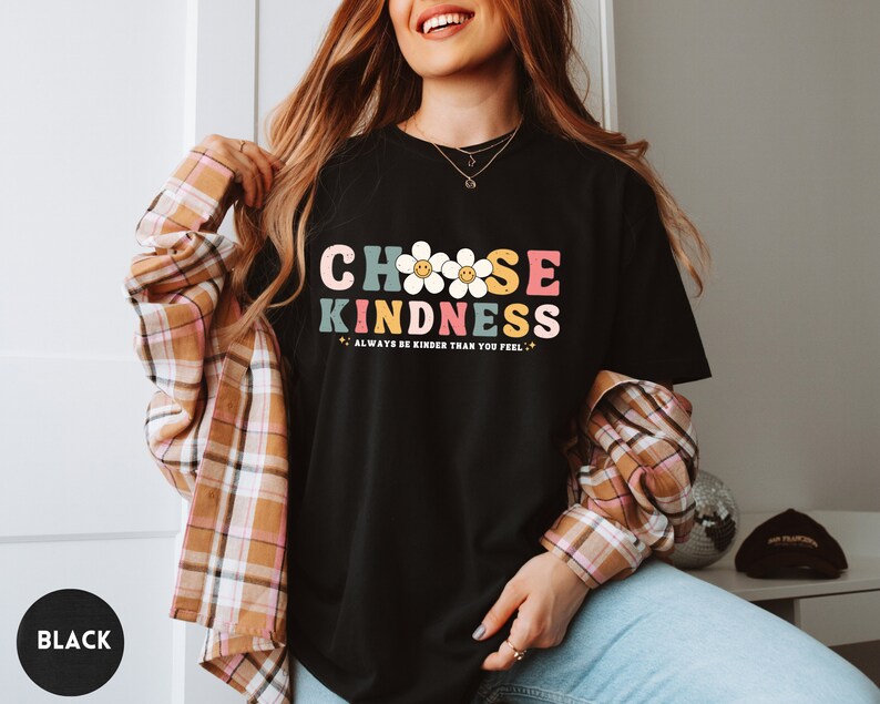 Peut inclure: T-shirt noir avec l'inscription "CHOOSE KINDNESS" dans un style r&eacute;tro color&eacute;. Le motif comprend des marguerites et la phrase "ALWAYS BE KINDER THAN YOU FEEL". Un cercle noir avec le mot "BLACK" est en bas &agrave; gauche.
