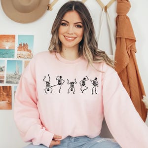 May include: A light pink sweatshirt featuring a black skeleton dance design. The graphic shows five dancing skeletons in various poses. The sweatshirt has long sleeves and a crew neck.