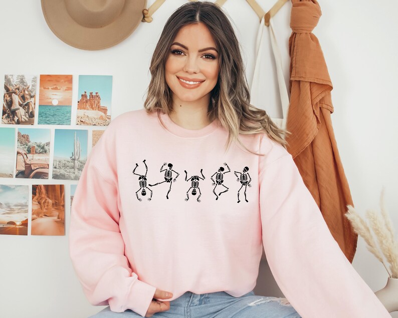 May include: A light pink sweatshirt featuring a black skeleton dance design. The graphic shows five dancing skeletons in various poses. The sweatshirt has long sleeves and a crew neck.