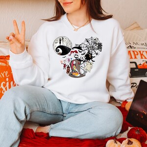 Puede incluir: Sudadera blanca de cuello redondo con un gr&aacute;fico de Jack Skellington y otros personajes de Pesadilla antes de Navidad. El dise&ntilde;o tiene forma de orejas de Mickey Mouse. La sudadera la lleva una persona.