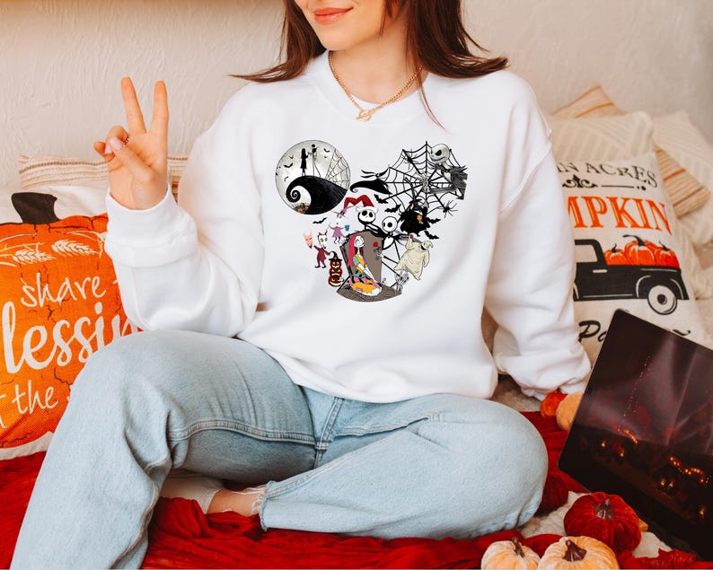 Puede incluir: Sudadera blanca de cuello redondo con un gr&aacute;fico de Jack Skellington y otros personajes de Pesadilla antes de Navidad. El dise&ntilde;o tiene forma de orejas de Mickey Mouse. La sudadera la lleva una persona.