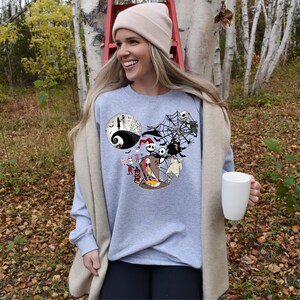 Puede incluir: Una sudadera gris claro con un gr&aacute;fico tem&aacute;tico de Halloween con personajes de Pesadilla antes de Navidad. El dise&ntilde;o incluye a Jack Skellington, Sally y otros personajes. La persona sostiene una taza blanca.