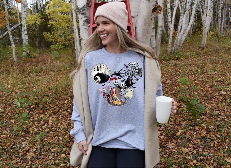 Puede incluir: Una sudadera gris claro con un gr&aacute;fico tem&aacute;tico de Halloween con personajes de Pesadilla antes de Navidad. El dise&ntilde;o incluye a Jack Skellington, Sally y otros personajes. La persona sostiene una taza blanca.