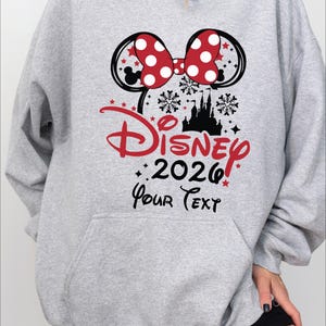 Puede incluir: Una sudadera con capucha gris claro con un gran gr&aacute;fico que presenta un lazo de lunares rojos y blancos, orejas de Mickey Mouse y la palabra "Disney" en rojo. El a&ntilde;o "2026" est&aacute; debajo, con espacio para texto personalizado.