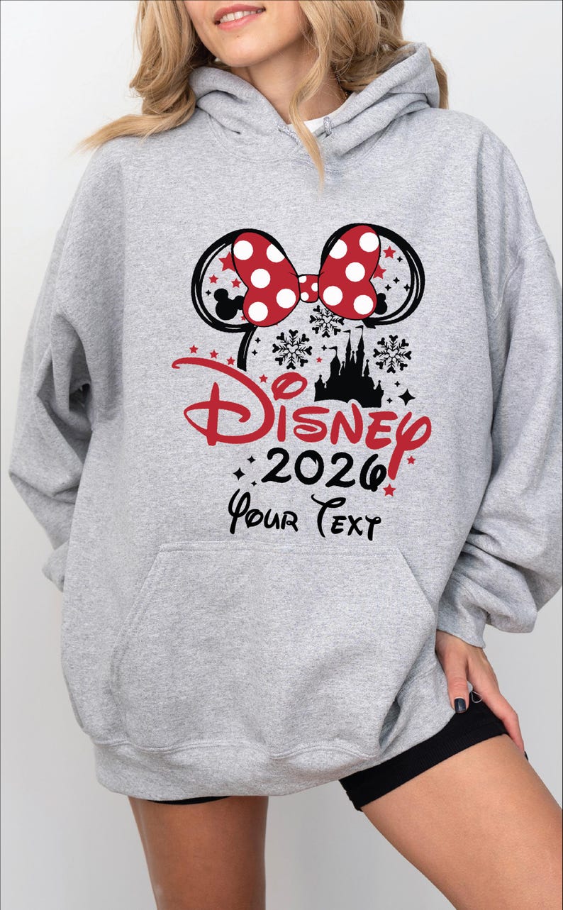 Puede incluir: Una sudadera con capucha gris claro con un gran gr&aacute;fico que presenta un lazo de lunares rojos y blancos, orejas de Mickey Mouse y la palabra "Disney" en rojo. El a&ntilde;o "2026" est&aacute; debajo, con espacio para texto personalizado.
