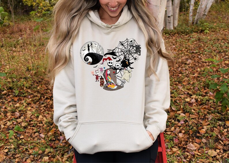 Puede incluir: Sudadera con capucha de color crema con un gr&aacute;fico de personajes de Pesadilla antes de Navidad. El dise&ntilde;o incluye a Jack Skellington, Sally y otros personajes, con una telara&ntilde;a y un fondo de luna. La sudadera tiene un bolsillo delantero y una capucha con cord&oacute;n.