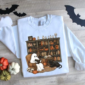 May include: White sweatshirt with a Halloween-themed graphic. The design features a ghost reading in a chair in front of a bookshelf, with pumpkins, candles, and a black cat. Decorative bats and pumpkins are also present.