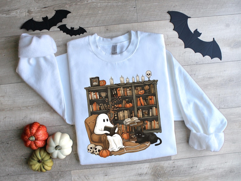 May include: White sweatshirt with a Halloween-themed graphic. The design features a ghost reading in a chair in front of a bookshelf, with pumpkins, candles, and a black cat. Decorative bats and pumpkins are also present.