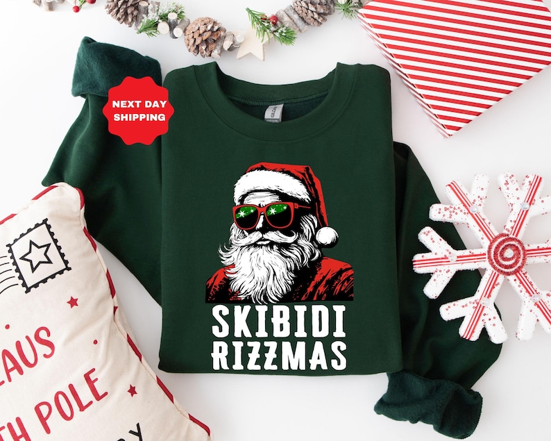 Puede incluir: Su&eacute;ter navide&ntilde;o verde con una imagen de dibujos animados de Pap&aacute; Noel con gafas de sol y un gorro rojo. El texto "Skibidi Rizzmas" est&aacute; impreso en blanco debajo de la imagen.