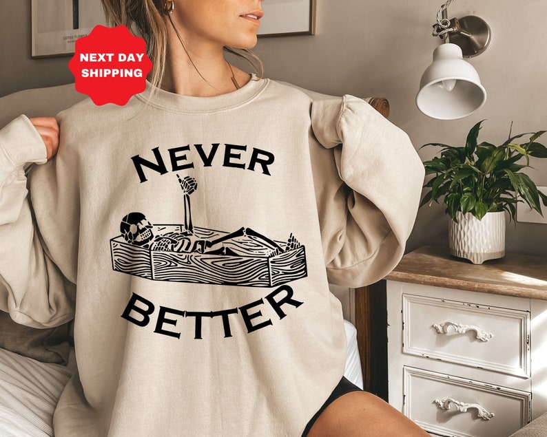 May include: A beige sweatshirt with a black graphic of a skeleton lying in a coffin with the text "NEVER BETTER" above it.