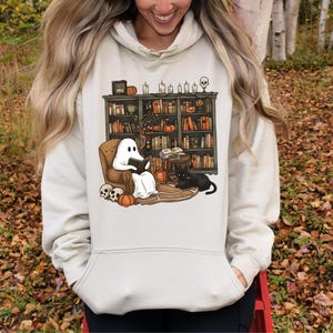 May include: A cream-colored hoodie featuring a Halloween-themed graphic. The graphic depicts a ghost reading in a chair in front of a bookshelf, with a black cat, pumpkins, and skulls. The design has a cozy, autumnal feel.