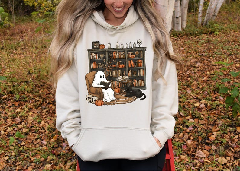 May include: A cream-colored hoodie featuring a Halloween-themed graphic. The graphic depicts a ghost reading in a chair in front of a bookshelf, with a black cat, pumpkins, and skulls. The design has a cozy, autumnal feel.