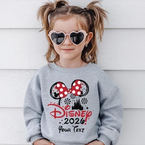 Puede incluir: Una joven con una sudadera gris con un dise&ntilde;o de Minnie Mouse. El dise&ntilde;o incluye un lazo rojo con lunares, orejas de rat&oacute;n negras y el texto "Disney 2026 Your Text" en rojo y negro. La persona tambi&eacute;n lleva gafas de sol en forma de coraz&oacute;n.
