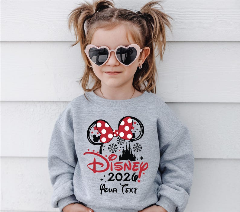 Puede incluir: Una joven con una sudadera gris con un dise&ntilde;o de Minnie Mouse. El dise&ntilde;o incluye un lazo rojo con lunares, orejas de rat&oacute;n negras y el texto "Disney 2026 Your Text" en rojo y negro. La persona tambi&eacute;n lleva gafas de sol en forma de coraz&oacute;n.