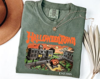 May include: A green t-shirt with an orange graphic of a spooky town with a large pumpkin in front of a building. The text "HALLOWEEN TOWN EST. 1998" is printed on the shirt.