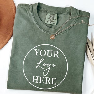 May include: A folded green t-shirt with a white circle that says "YOUR Logo HERE" on the front. The t-shirt is on a white surface with a brown hat and a dried palm leaf.