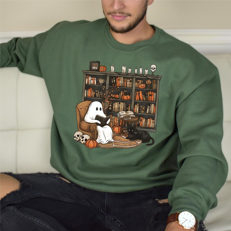May include: Green sweatshirt featuring a Halloween-themed graphic. The design shows a ghost reading in a chair in front of a bookshelf, with pumpkins, a black cat, and skulls. The sweatshirt is a crew neck style.