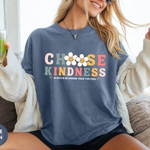 Peut inclure: T-shirt bleu jean avec l'inscription "CHOOSE KINDNESS" en lettres color&eacute;es de style r&eacute;tro. Le t-shirt pr&eacute;sente &eacute;galement des graphiques de marguerites et la phrase "ALWAYS BE KINDER THAN YOU FEEL". Fait partie d'une collection de v&ecirc;tements d&eacute;contract&eacute;s.