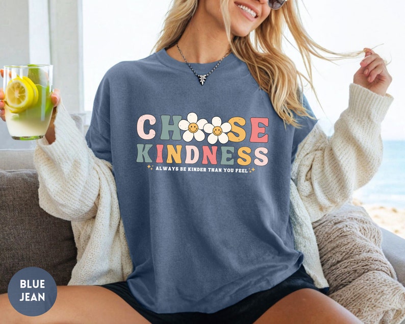 Peut inclure: T-shirt bleu jean avec l'inscription "CHOOSE KINDNESS" en lettres color&eacute;es de style r&eacute;tro. Le t-shirt pr&eacute;sente &eacute;galement des graphiques de marguerites et la phrase "ALWAYS BE KINDER THAN YOU FEEL". Fait partie d'une collection de v&ecirc;tements d&eacute;contract&eacute;s.