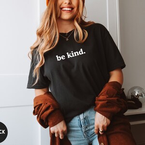 Op de afbeelding: Een zwart T-shirt met de tekst "be kind." in wit gedrukt op de voorkant. Het shirt wordt gedragen door een persoon met lang blond haar en een bruin jack.