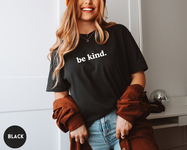 Op de afbeelding: Een zwart T-shirt met de tekst "be kind." in wit gedrukt op de voorkant. Het shirt wordt gedragen door een persoon met lang blond haar en een bruin jack.