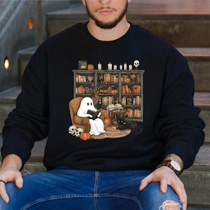 May include: Black sweatshirt featuring a Halloween-themed illustration. A ghost reads in a cozy armchair in front of a bookshelf filled with books, pumpkins, and candles. A black cat sits nearby. Skulls and pumpkins are also present.